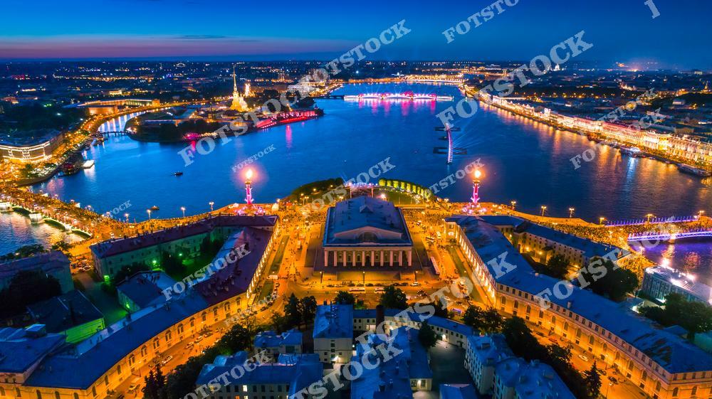 На фотографии запечатлена Стрелка Васильевского острова, украшенная для праздника Алые Паруса, который ежегодно проводится в Санкт-Петербурге. Яркие огни Ростральных колонн, здания Биржи и других исторических построек создают волшебную атмосферу праздника. Вдали виднеются красные паруса на корабле, символизирующие событие, а огни набережных отражаются в спокойных водах Невы. Этот вид является символом красоты и величия Петербурга в ночное время.